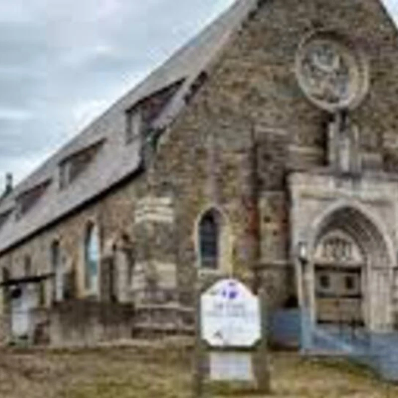 A low-angle shot of a large, weathered stone church with Gothic-style arched windows and a prominent rose window. The sky above is gray and cloudy.